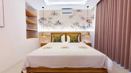 Cozy tropical bedroom featuring a wooden king bed with white linens, palm leaf wallpaper, rattan pendant lights, and soft pink curtains.