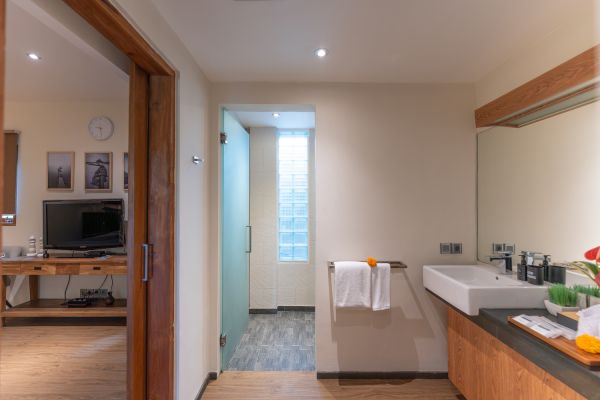 Modern en-suite hotel bathroom with wooden vanity, white sink, hanging towels, and open doorway to adjacent bedroom with TV and desk.
