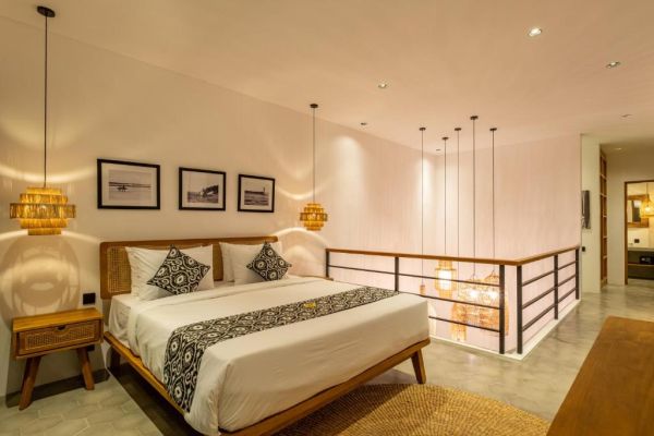 Cozy Balinese-style loft bedroom with wooden bed, batik bedding, rattan pendant lamps, and overlooking railing.