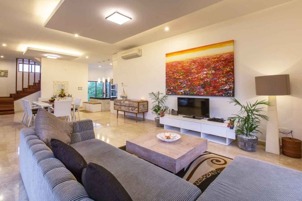 Spacious modern Balinese living room with gray sectional sofa, abstract floral wall art, wooden dining set, and potted plants. - Bali Villas