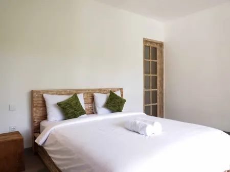 Cozy minimalist bedroom with white duvet, green pillows, rolled towel, and wooden bed frame against white walls.