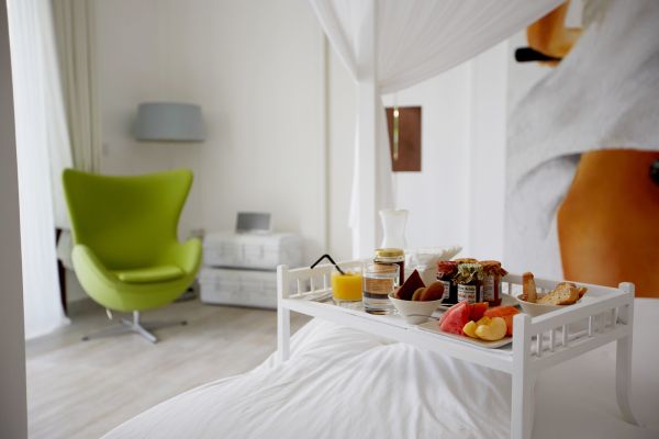 Luxurious bedroom with breakfast tray on white bed featuring orange juice, fresh fruits, pastries, and jams, beside a lime-green armchair and sheer curtains.
