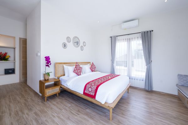Cozy Balinese-style bedroom with white wooden bed, red embroidered runner, orchids, and sheer-curtained windows.