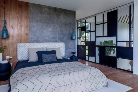 Modern bedroom interior with wooden accent wall, gray patterned bedding, blue pendant lights, and black-framed sliding doors overlooking greenery.