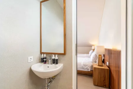 Modern minimalist bathroom sink and mirror beside an open door to a cozy bedroom with twin white beds and wooden nightstands.