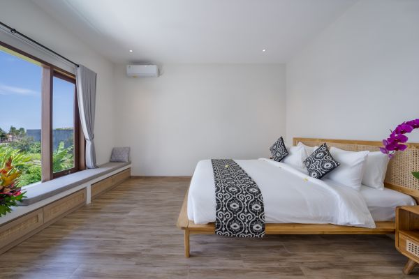 Spacious tropical bedroom with wooden bed, white linens, black patterned accents, orchids, and panoramic window view of lush Balinese greenery.