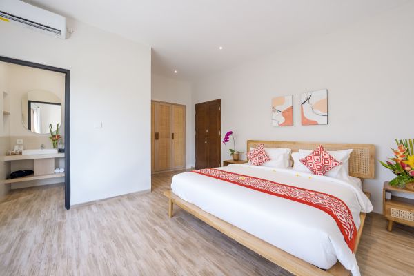 Spacious Balinese bedroom with white linens, red patterned accents, tropical flowers, abstract wall art, and open en-suite bathroom.