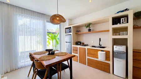 Bright modern tropical kitchen with wooden cabinets, dining table, rattan pendant light, plants, and sheer-curtained windows. - Bali Villas