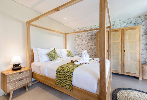 Elegant tropical bedroom with wooden four-poster bed, white linens, green patterned accents, swan towel decoration, floral wallpaper, and rattan wardrobe.