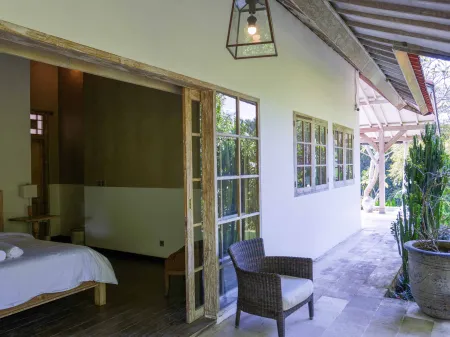 Serene Balinese villa bedroom with white bed, opening to tropical veranda featuring rattan chair, hanging lantern, and potted plants.