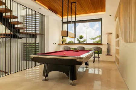 Modern game room featuring a sleek black pool table with red felt, surrounded by wooden stools, under a wooden ceiling with pendant lights, large windows showing tropical greenery, and stylish wooden accents. - Bali Villas