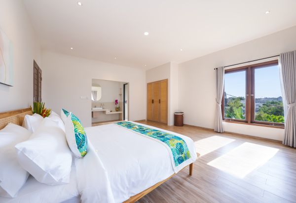 Spacious Bali villa bedroom with wooden bed, tropical green bedding, en-suite bathroom, and lush garden view through large windows.