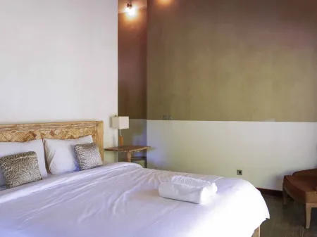 Cozy tropical bedroom with carved wooden bed, white linens, folded towel, and ambient wall lighting.
