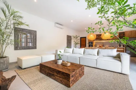Modern tropical living room interior with white L-shaped sofa, wooden coffee table, potted plants, hanging lanterns, and open kitchen. - Bali Villas