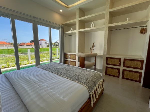 Spacious tropical bedroom with king bed, rattan furniture, and panoramic view of green rice fields through floor-to-ceiling windows.