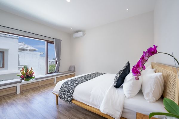 Serene tropical bedroom interior with wooden bed, white linens, purple orchids, and large window overlooking lush greenery.