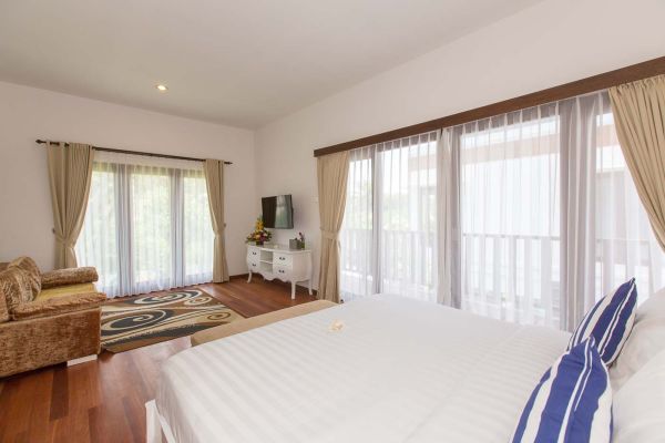 Spacious tropical bedroom with king bed, cream sofa, wall-mounted TV, and balcony views through sheer-curtained windows.