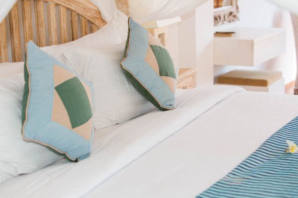 Close-up of a luxurious white bed with blue-and-green quilted pillows and a frangipani flower in a tropical bedroom.