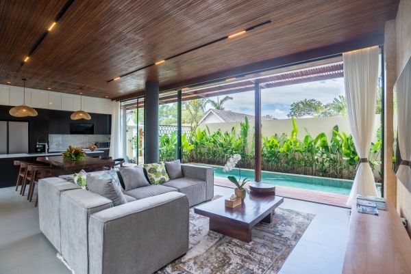 Modern tropical living room with gray sofa, wooden accents, open kitchen, and sliding glass doors to lush garden pool. - Bali Villas