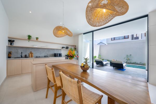 Modern tropical kitchen and dining area with wooden table, rattan chairs, hanging lamps, and infinity pool view through glass doors. - Bali Villas