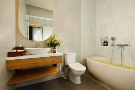 Modern spa bathroom featuring wooden vanity with round sink, oval mirror, white toilet, and petal-filled freestanding tub. - Bali Villas