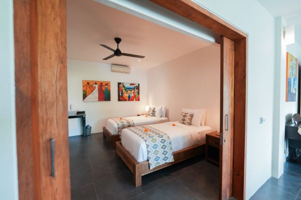 Spacious Balinese hotel bedroom with two queen beds, wooden accents, ceiling fan, and vibrant wall art, framed by open sliding doors.