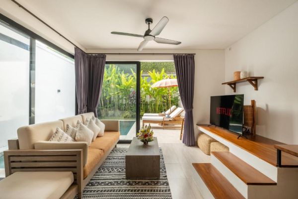 Modern tropical villa interior with beige sofa, wooden accents, Netflix TV, and glass doors to private pool lounge. - Bali Villas