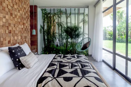 Modern tropical bedroom with wooden accent wall, lush indoor plants, king bed in white linens and black geometric throw, hanging green pendant, wicker egg chair, and garden view through floor-to-ceiling glass doors.