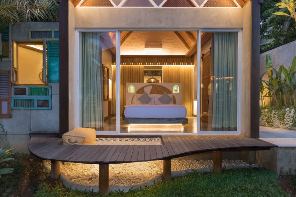 Luxurious tropical bedroom in modern villa with open glass doors revealing king bed on wooden deck amid lush garden at dusk.