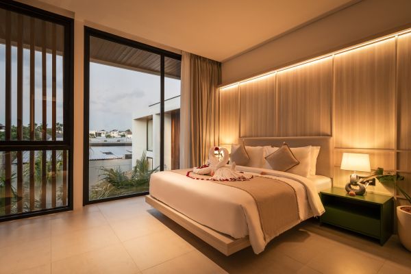 Luxurious modern bedroom with king bed decorated in rose petals, floor-to-ceiling windows overlooking tropical water and villas, warm ambient lighting.
