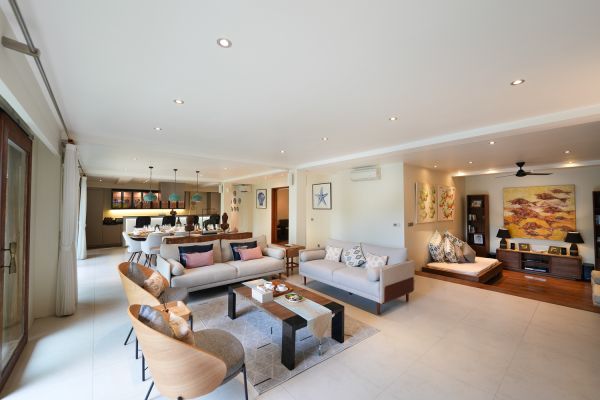 Spacious modern living room with light beige sofas, wooden chairs, abstract wall art, and open kitchen in a tropical-style home. - Bali Villas