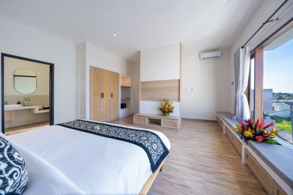 Spacious Balinese bedroom with king bed, wooden accents, tropical flowers, ensuite bath, and lush green window view.