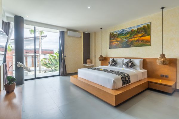 Spacious Bali villa bedroom with wooden platform bed, white linens, tropical palm views through floor-to-ceiling windows, and landscape painting.
