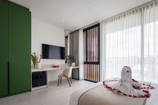 Luxurious modern hotel bedroom with king bed topped by towel swan and rose petals, green wardrobe, desk with plants, wall TV, and large sheer-curtained windows.
