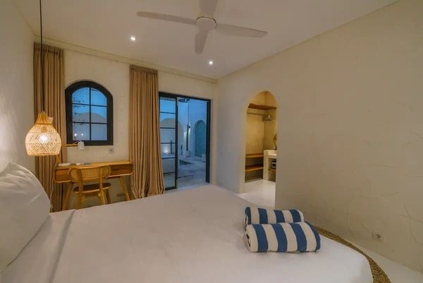 Cozy tropical bedroom with white bed topped by striped towels, wooden desk, ceiling fan, and arched doors to a courtyard at dusk.