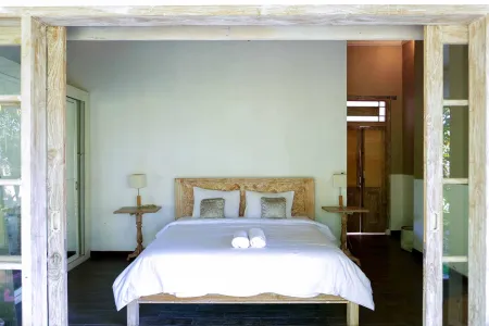 Serene tropical bedroom with wooden king bed, white linens, folded towels, and open doors revealing lush greenery. - Bali Villas