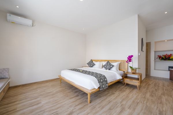 Serene minimalist bedroom with wooden platform bed, white linens, orchids on nightstand, and tropical decor in a light-filled villa.