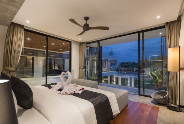 Luxurious tropical bedroom with romantic towel swans and rose petals on king bed, floor-to-ceiling windows overlooking infinity pool at sunset. - Bali Villas