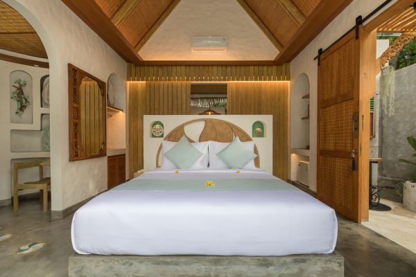 Luxurious Balinese villa bedroom with white king bed, frangipani accents, wooden headboard, high beamed ceiling, and garden view through arched sliding doors. - Bali Villas