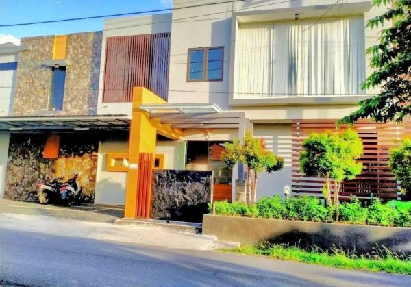 Modern Indonesian house exterior with white walls, orange accents, stone and wood features, parked scooter, and tropical plants on a sunny street. - Bali Villas