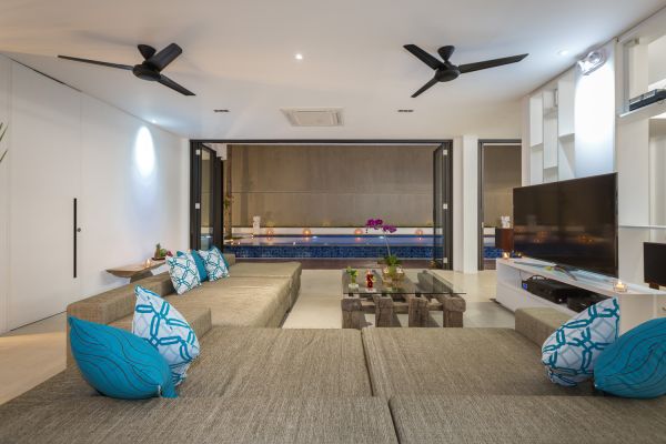Modern open-plan living room in a tropical villa, featuring low beige sofas with blue pillows, wooden coffee table with drinks and orchids, and sliding doors to an infinity pool. - Bali Villas