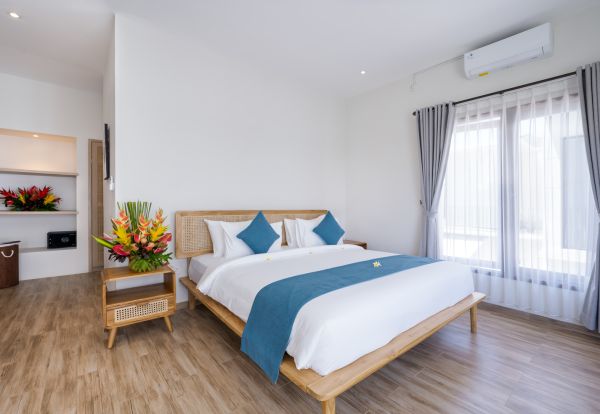 Serene Balinese-style bedroom with wooden platform bed, white linens, blue cushions, tropical flower vase, and large curtained windows.