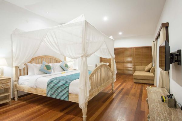 Spacious Balinese-style bedroom with white canopy four-poster bed, turquoise bedding, wooden floors, and wall-mounted TV.