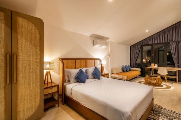Cozy tropical bedroom interior featuring a wooden platform bed with blue pillows, rattan wardrobe, orange sofa, and high vaulted ceiling with dark curtains. - Bali Villas