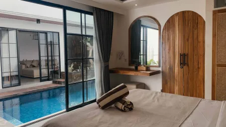 Modern tropical bedroom with large glass doors overlooking a private blue infinity pool, wooden vanity, and neatly made bed.