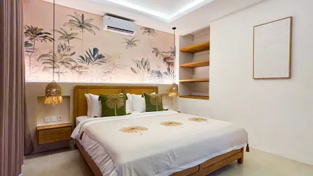 Cozy modern tropical bedroom featuring palm leaf wallpaper, rattan pendant lights, wooden bed with white linens, and built-in shelves. - Bali Villas