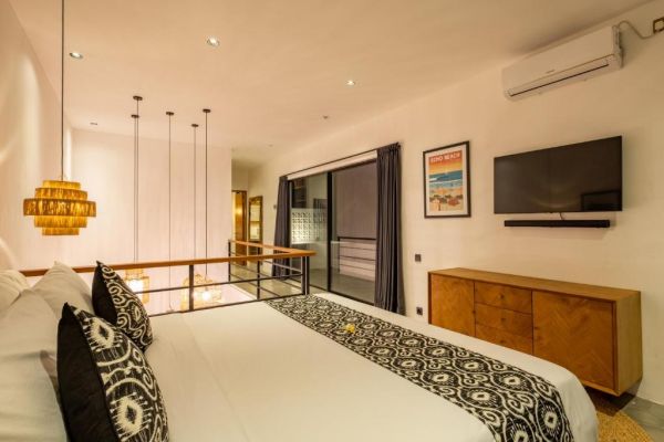 Modern Bali villa bedroom with king bed, patterned linens, wall-mounted TV, sliding balcony doors, and rattan pendant lights.