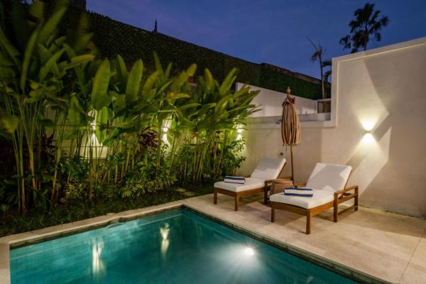 Serene tropical infinity pool at dusk with white lounge chairs, beige umbrella, and lush banana palms under soft lighting. - Bali Villas