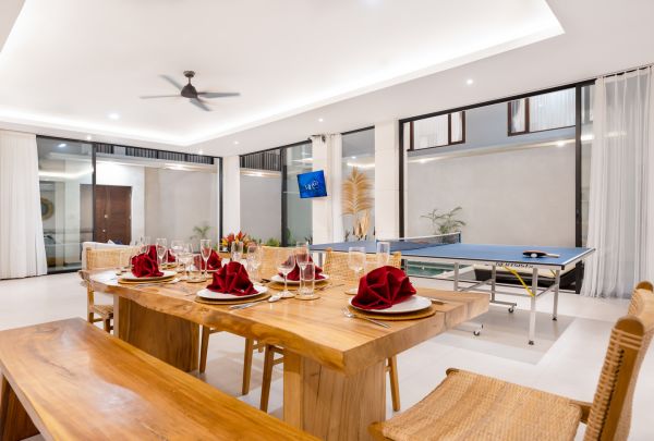 Spacious tropical dining room with wooden table set for meal, red napkins, and nearby ping pong table, large glass windows, ceiling fan, and pool view. - Bali Villas