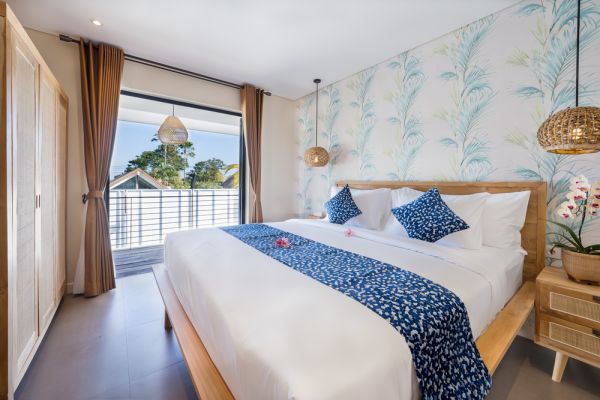 Cozy Balinese bedroom with white wooden bed, blue patterned runner, rattan lights, leaf wallpaper, orchids, and tropical balcony view.